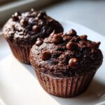Close-up of two moist chocolate zucchini muffins topped generously with chocolate chips, sitting on a white plate.