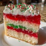 A festive slice of christmas poke cake showing layers of white cake, red and green jello, whipped cream, and sprinkles.