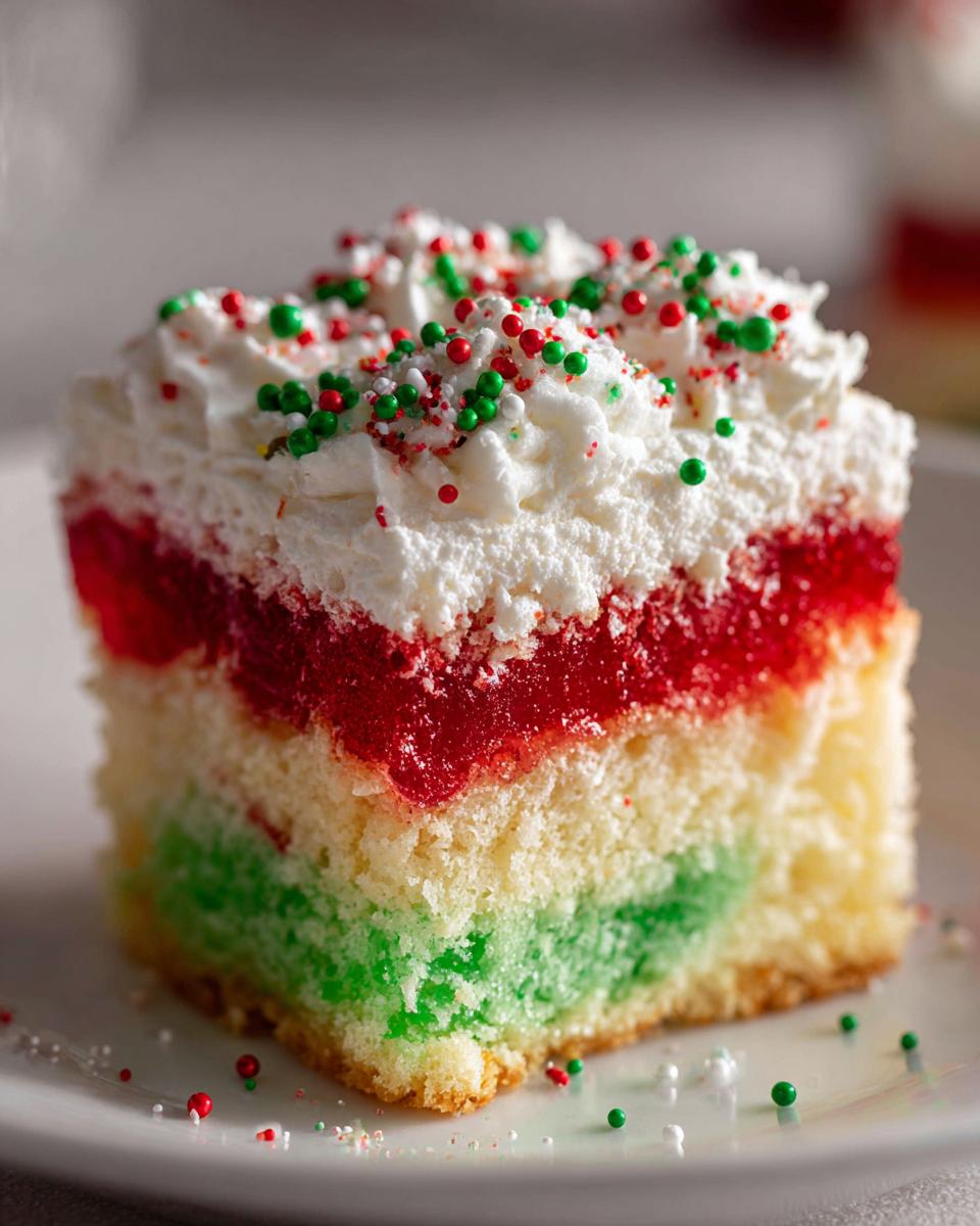Close-up of a slice of Christmas poke cake showing layers of green cake, red gelatin, whipped topping, and festive sprinkles.