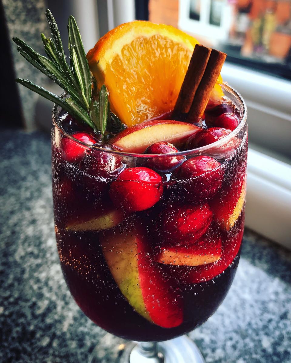 Close-up of a glass filled with sparkling Christmas sangria, garnished with cranberries, orange slice, cinnamon sticks, and rosemary.