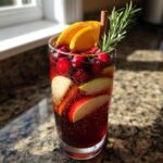 Close-up of a tall glass filled with bubbly Christmas sangria, garnished with cranberries, orange slices, apple, rosemary, and a cinnamon stick.