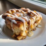 A close-up of a rich cinnamon roll apple pie slice topped with thick white icing, sitting on a white plate.
