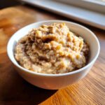 Close-up of creamy banana oatmeal topped with cinnamon served in a white bowl on a wooden surface.