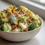 A close-up view of a white bowl filled with creamy broccoli salad, topped generously with crispy bacon bits and shredded cheddar cheese.