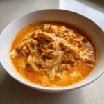 Close-up of a white bowl filled with creamy, orange-hued buffalo chicken chili topped with shredded chicken and melted cheese.