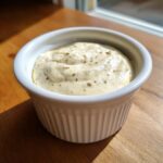 A small white ramekin filled with creamy, pale yellow horseradish sauce speckled with dark spices.