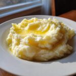 A close-up of fluffy, creamy mashed potatoes topped with melted butter, served on a white plate.