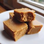 Close-up of several thick squares of creamy peanut butter fudge stacked on a white plate.