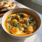 A close-up of a creamy, orange pumpkin curry topped with fresh cilantro and a swirl of cream, served with naan bread in the background.