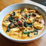 A close-up, appetizing view of creamy sausage tortellini soup filled with pasta, ground sausage, and wilted spinach.