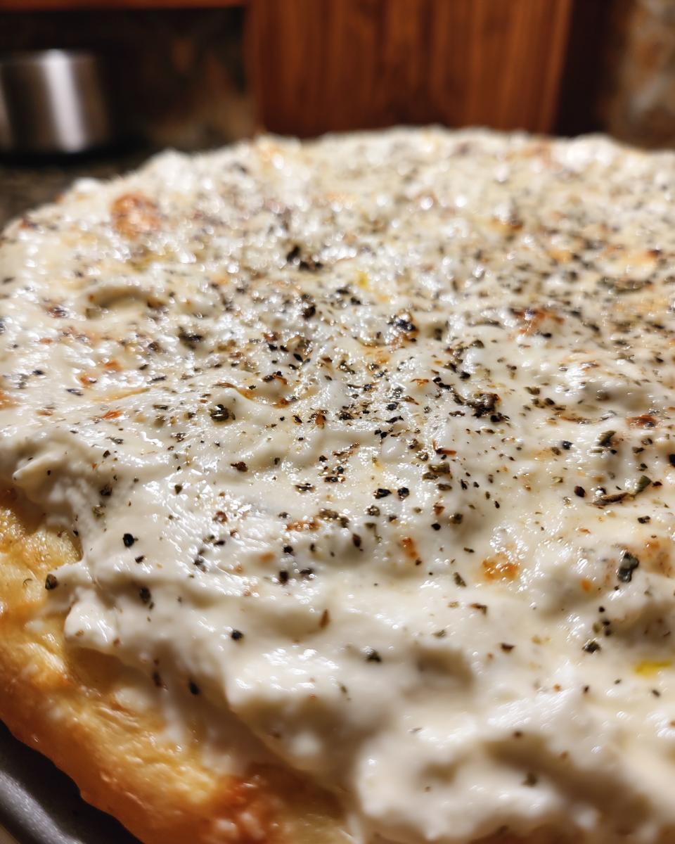 Close-up of a golden crust topped generously with creamy white pizza sauce and sprinkled with black pepper.