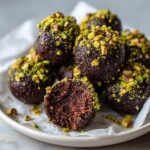 Close-up of rich, dark Dubai chocolate balls rolled in chopped pistachios, one cut in half showing the dense center.