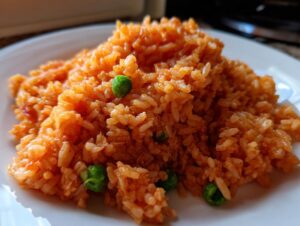 A close-up, featured image of a mound of bright orange, fluffy spanish rice mixed with a few bright green peas, served on a white plate.