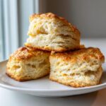 Three fluffy, golden-brown scones stacked on a white plate, showing off their flaky layers.