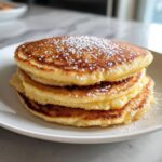 A tall stack of three fluffy coconut flour pancakes dusted generously with white powdered sugar on a white plate.