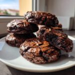 A stack of fudgy chocolate brownie cookies with shiny, cracked tops, one broken open to show the dense interior.