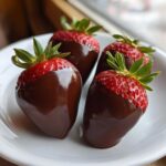 Four perfectly dipped, glossy chocolate covered strawberries resting on a white plate.