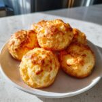 A small plate holding five freshly baked, golden brown brazilian cheese bread puffs with crispy tops.