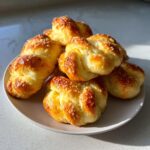 A stack of golden brown, freshly baked garlic parmesan knots topped with melted cheese, sitting on a white plate.