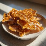 Close-up of broken, shiny pieces of homemade peanut brittle recipe served on a small, light-colored plate.