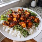 Close-up of a bowl filled with white rice, topped with glazed harissa chicken pieces, arugula, and sesame seeds.