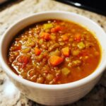 Close-up of a white bowl filled with thick, hearty lentil soup, showing visible carrots and celery pieces.
