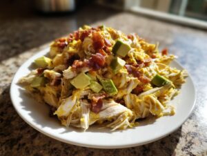 A mound of shredded chicken coated in yellow honey mustard dressing, topped with crispy bacon bits and diced avocado, served on a white plate.
