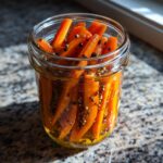 Close-up of bright orange pickled carrots spears packed in a glass jar with peppercorns and pickling spices.