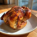 A beautifully roasted whole chicken with dark, crispy, herb-seasoned skin resting on a white plate.