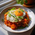 A vibrant plate of kimchi fried rice topped with a sunny-side-up egg, sesame seeds, and sliced green onions.