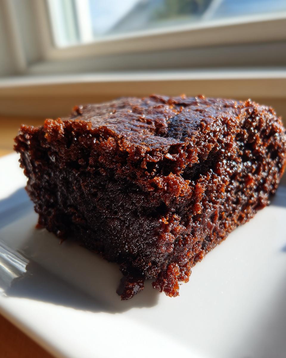 Close-up of a rich, dark, and moist square of date brownies sitting on a white plate near a window.
