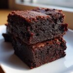 Close-up of two stacked, rich, dark chocolate date brownies on a white plate.