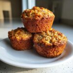 Three golden brown oatmeal muffins stacked on a white plate, featuring visible rolled oats on top.