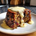 Close-up of a rich, dark slice of plum pudding studded with dried fruit and nuts, generously covered in creamy custard.