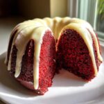 A moist red velvet bundt cake, cut in half to show the rich red crumb, topped with dripping white glaze.