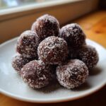 A close-up stack of rich, dark bourbon balls generously coated in shredded coconut resting on a white plate.