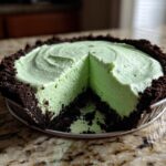 A close-up of a no-bake grasshopper pie with a thick, light green filling and dark chocolate crust, showing one slice removed.