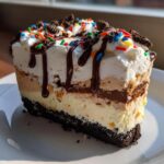 A rich slice of no-bake ice cream cake showing multiple layers, topped with whipped cream, chocolate syrup, and sprinkles.
