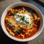 Close-up of a white bowl filled with rich, red one pot lasagna soup, topped with melted cheese, a dollop of ricotta, and parsley.