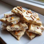 A stack of broken pieces of white chocolate peanut butter pretzel bark topped with nuts and small pretzels.