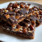 Close-up of stacked squares of rich Pecan Pie Bark topped with pecans, caramel, and dark chocolate chunks.