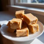 A stack of creamy, light brown penuche fudge squares resting on a white dish near a window.