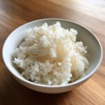 Close-up of perfectly cooked, glossy sushi rice served in a small white bowl on a wooden surface.