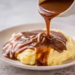 Thick, rich homemade brown gravy being poured from a white gravy boat onto a mound of creamy mashed potatoes.