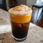A tall glass of pumpkin cream cold brew with distinct layers of dark coffee, orange pumpkin foam, and white whipped topping dusted with cinnamon.