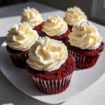 A close-up of several vibrant red velvet cupcakes topped with swirls of cream cheese frosting.