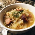 Close-up of a white bowl filled with rich sauerkraut soup, featuring chunks of smoked sausage and fresh dill garnish.