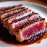 Close-up of perfectly seared Tuna steak, sliced and resting in a dark, rich sauce on a white plate.