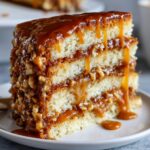 Close-up of a thick slice of caramel crunch cake showing three layers of sponge, caramel filling, and nuts, topped with dripping caramel sauce.