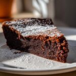 A close-up slice of rich, dark flourless chocolate cake dusted generously with white powdered sugar on a white plate.